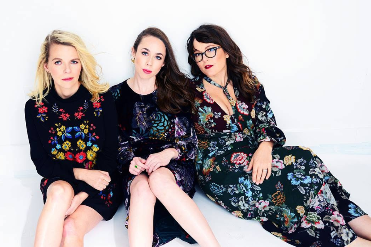 Three people wearing dresses sitting in front of a white backdrop.