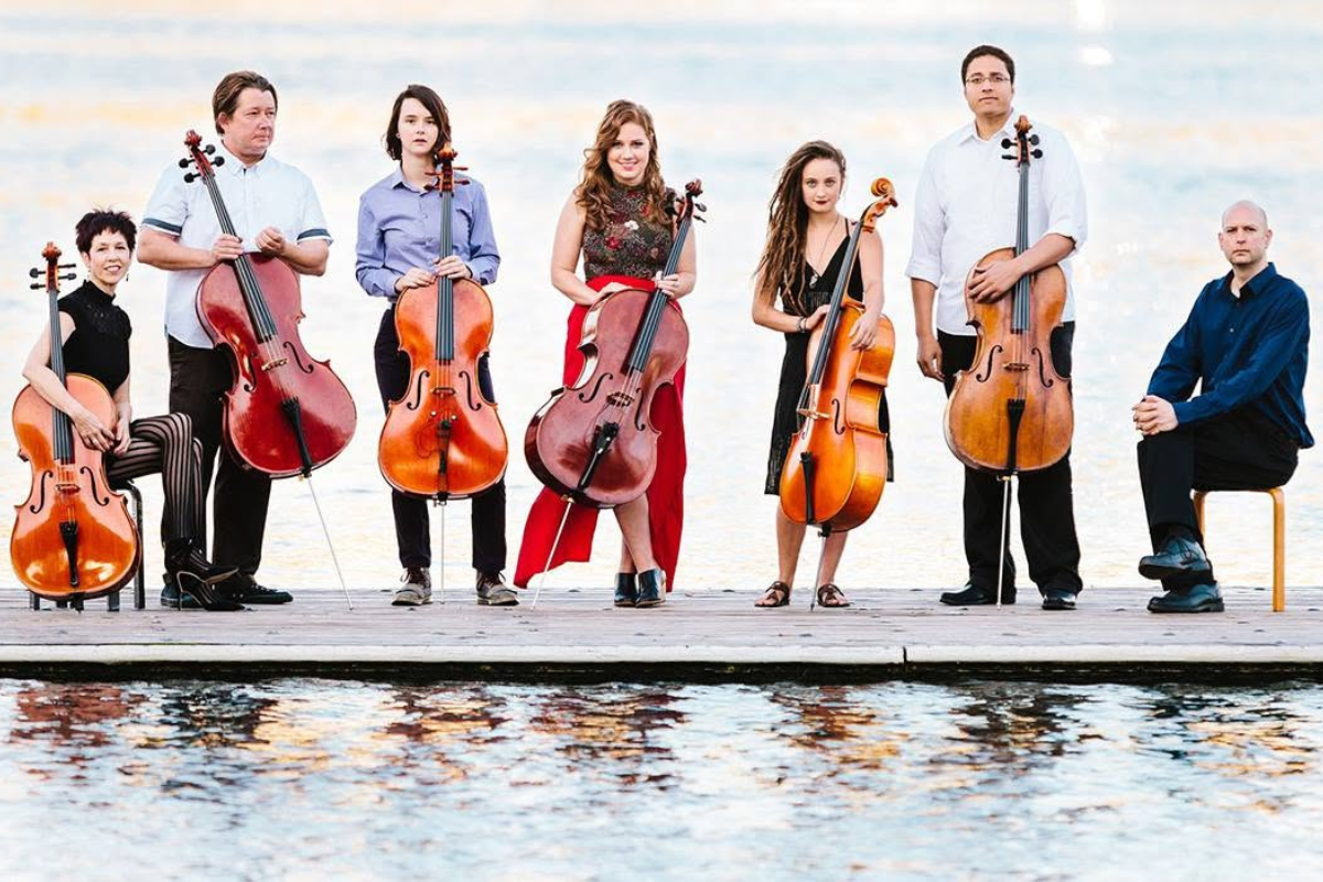 A group of cellists in a line, standing on a dock.