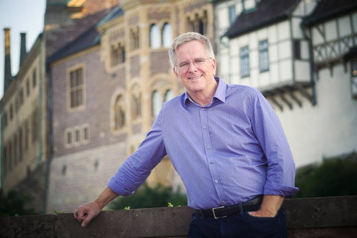 Photo of a person wearing a purple dress shirt, leaning on a railing in front of buildings.