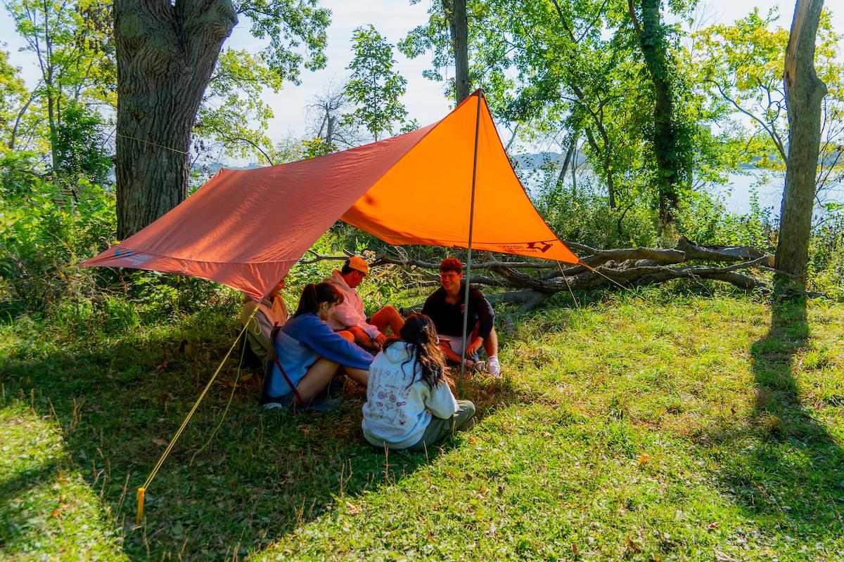 Four people sit in the grass under an orange tent
