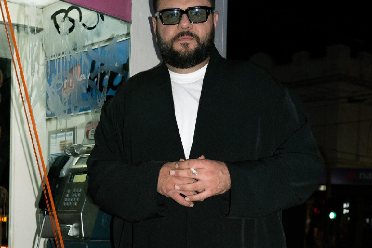 Photo of Mo Amer dressed in black, standing against a phone booth.