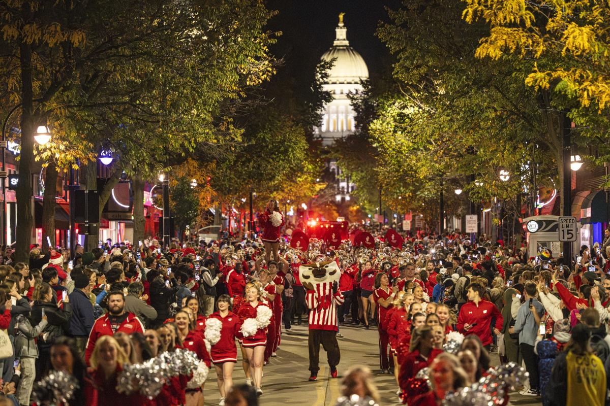 Bucky Badger struts down State Street with cheerleaders and marching band members as audience members cheer them on from the parade sidelines.