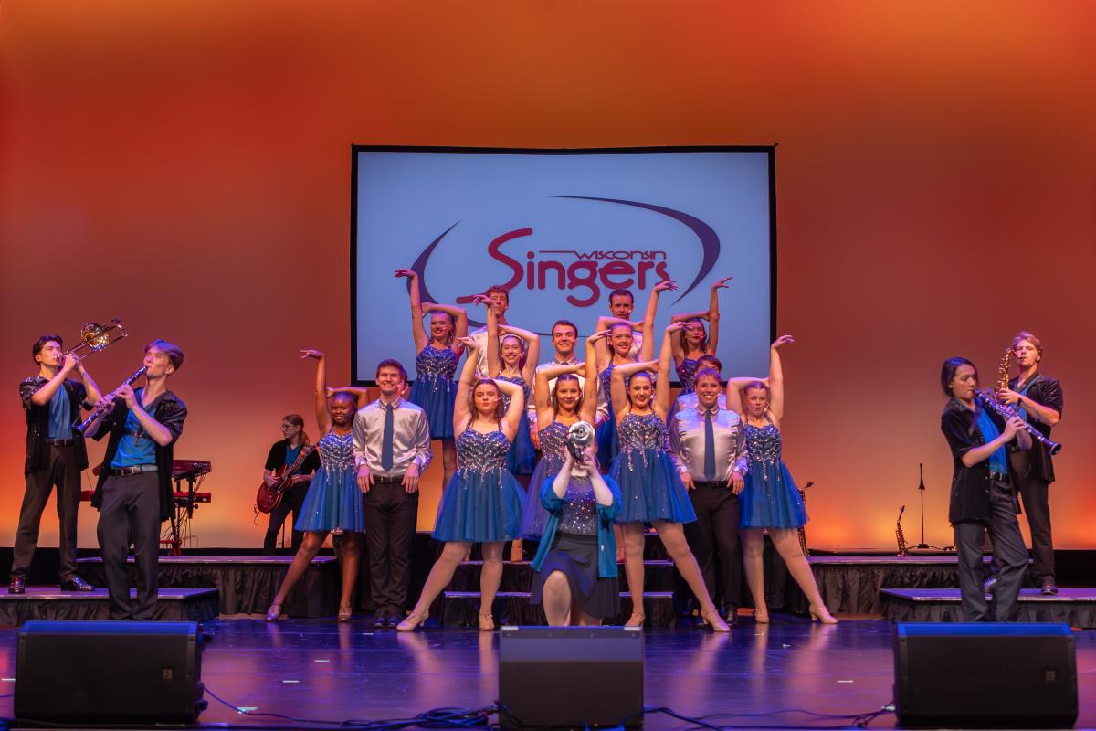 A group of performers in blue shimmery dresses and collared shirts and ties pose in front of a banner that reads &quot;Wisconsin Singers&quot; while band members play various instruments around them.
