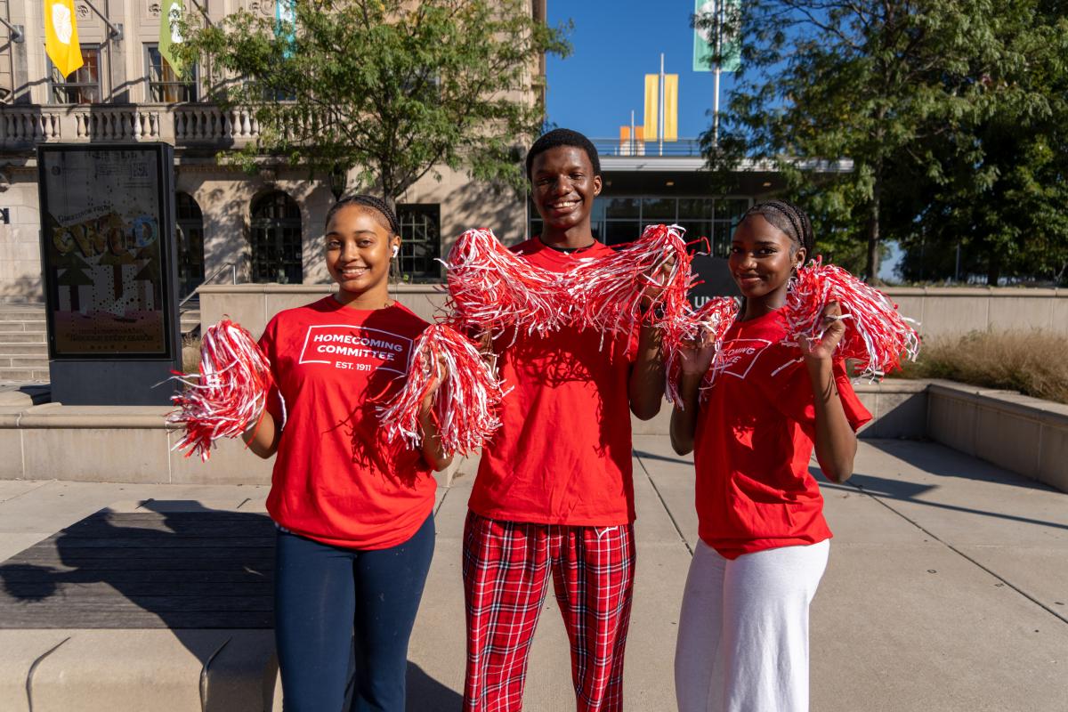 Three UW Homecoming Committee members standing and smiling with pom poms in their hands