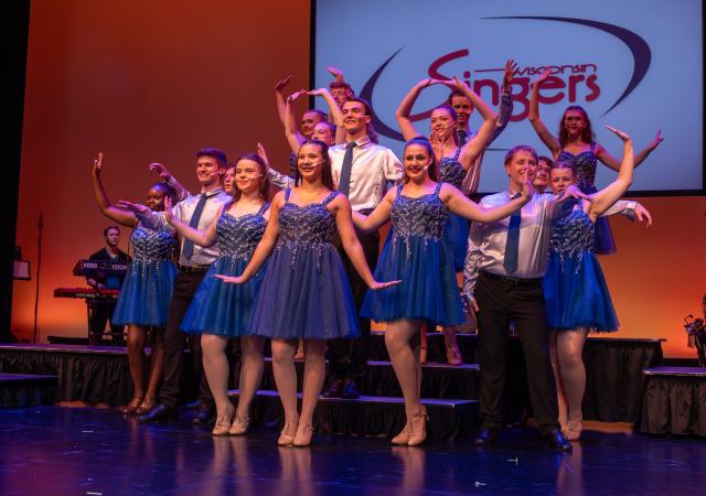 Wisconsin Singers pose on the stairs wearing blue sparkly dresses and silver shirts with an orange backdrop behind them.