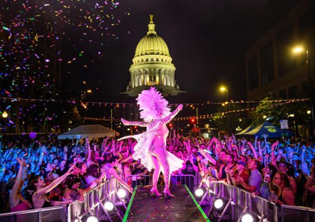 Drag Performer Cass Marie Domino performs on a stage near the Wisconsin State Capitol building at the Live on Queen Festival. She is lit brightly in purple and pink, and there are many excited fans with their hands raised above their heads. The Wisconsin State Capitol Building is well lit in the background.