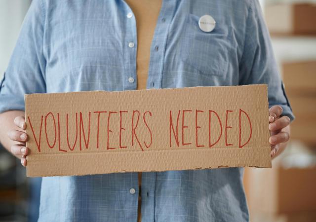 torso of an individual holding a cardboard sign reading &quot;volunteers needed&quot; written in red ink