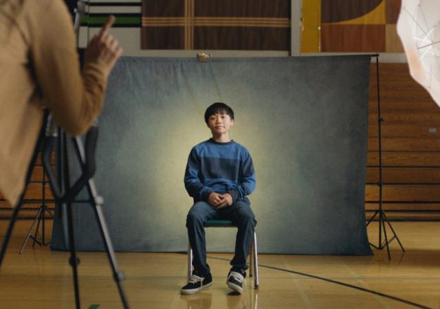 Still from film Didi, kid sits in front of grey background for school picture day.