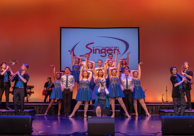 A group of performers in blue shimmery dresses and collared shirts and ties pose in front of a banner that reads "Wisconsin Singers" while band members play various instruments around them.
