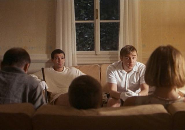 Still from film Funny Games (1997), family sits on couch and home invaders sit across from them.