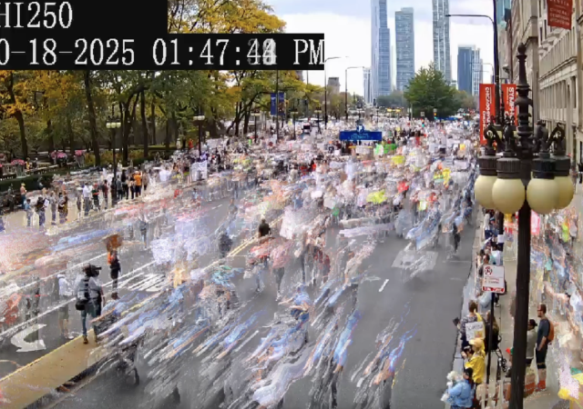 An image from a camera looking at a busy street in a non-descript city. There is a timestamp in white text over a black box in the upper left-hand corner. People are moving quickly and are blurred due to the motion. The image is slightly grainy as it is taken from a surveillance camera. There are trees on the opposite side of the street, and buildings and lampposts on the side of the street the camera is on.a