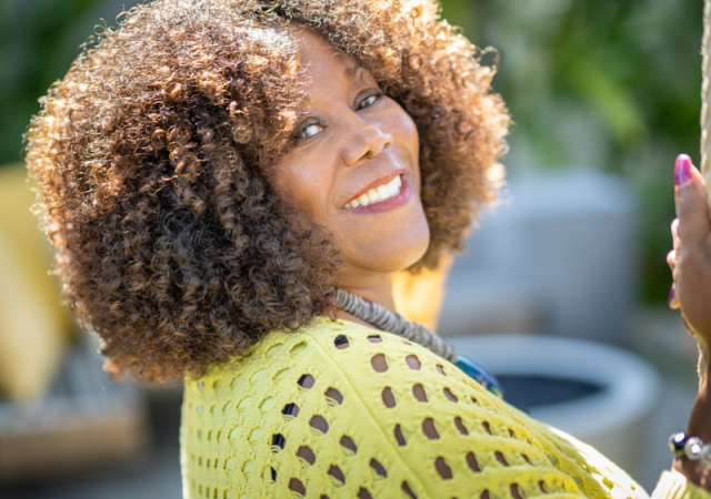 A smiling woman with curly hair wearing a bright yellow sweater looks back over her shoulder while sitting on a swing outdoors in a sunny garden.