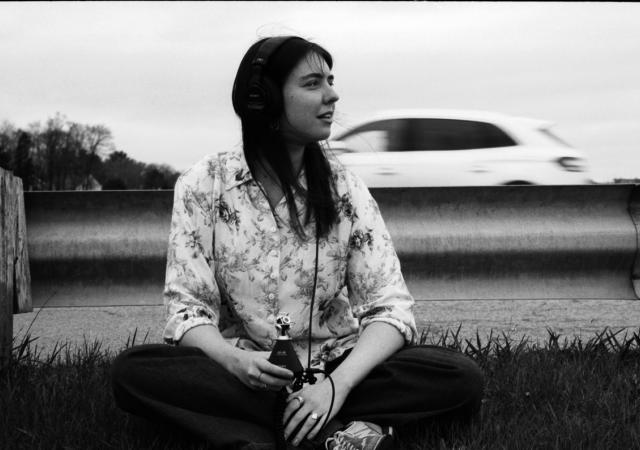 A black-and-white image of artist Anne E. Stoner. She is sitting down near the side of a road, with cars in motion behind the guardrail behind her. She is holding a microphone to record the sounds of traffic. She is looking to the right side of the image. She has long dark hair and is wearing light-colored shirt with a light pattern.