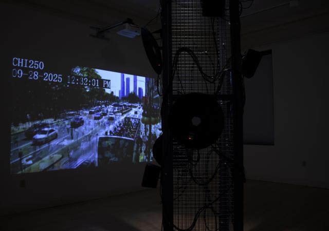 A dark image from inside Anne Stoner's Infrastructure Bodies/Injury Systems. In the foreground there is a metal structure with various speaker and wires strewn about. Behind the structure on the wall is a projected image of a streetscape with people and cars, the image is "datamoshed" meaning that pixels are merging into one another. The windows of Main Gallery are slightly visible in the background.