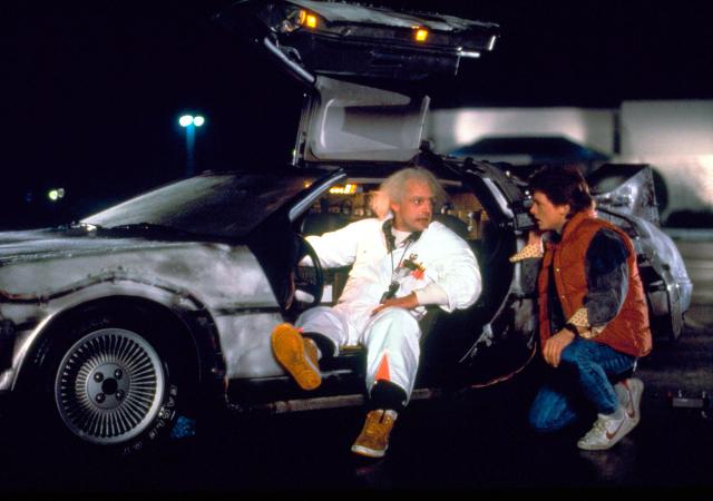 two individuals, one seated in a DeLorean vehicle and one crouching next to the vehicle