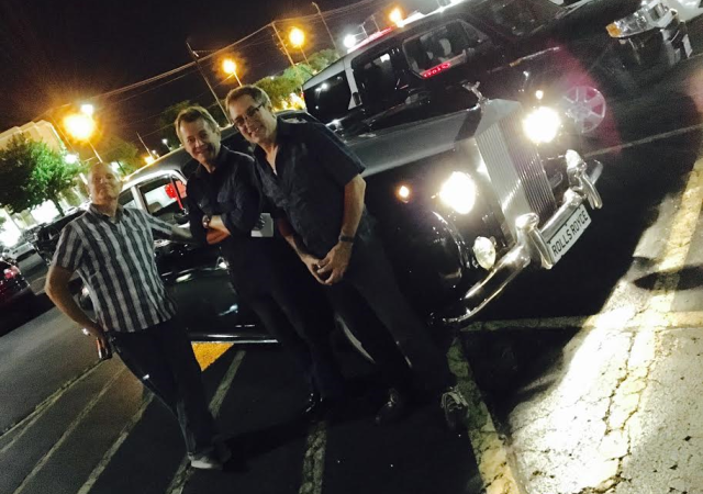 Three men stand in front of an old car, smiling for the camera.