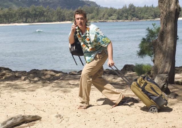 man dragging suitcase across beach with a body of water behind him