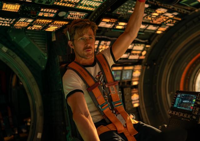 astronaut seated in space station seat wearing orange harness with lighted panel behind him