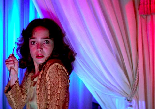 woman stands in a curtain-covered room holding a knife looking shocked and scared