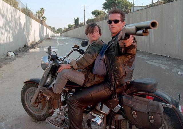 Two individuals sit on a motorcycle one points a gun behind the camera looking stoic.
