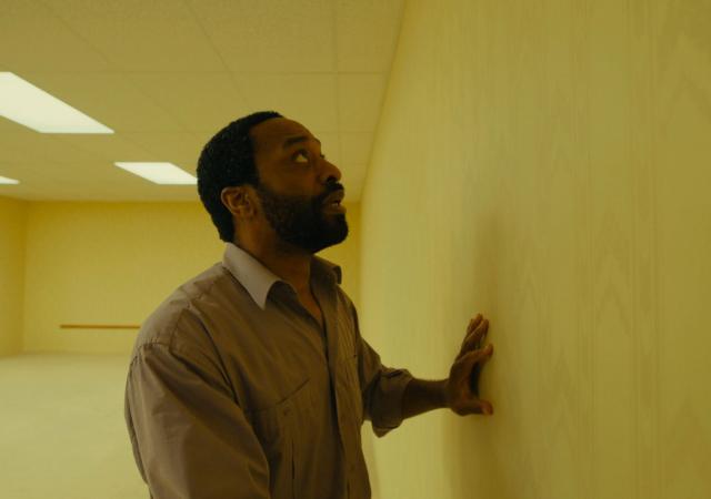Individual in a yellow, empty room, places his hand on a wall looking at it curiously and afraid.