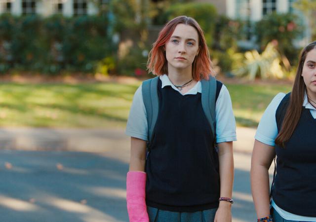two individuals walk down a street wearing dull school uniforms, looking sad, one has a hot pink cast on