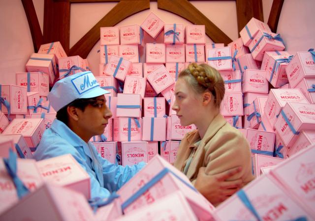 Two individuals sit in a truck full of pink cake boxes looking at eachother
