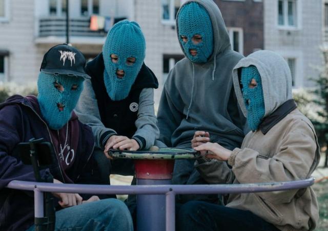 Four young individuals wearing blue ski masks sitting on a playground merry-go-round
