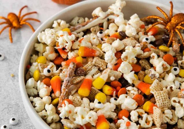 Halloween snack mix with popcorn, candy corn, and pretzels, and spiders and pumpkins on counter in background.