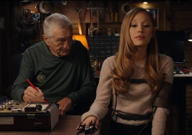 Two individuals sitting down in a dim room. One is strapped to a lie detector machine and the other is examining them and writing on a piece of paper