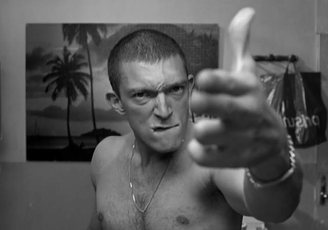 A shirtless man with a buzz cut and a chain necklace glares at the camera while extending his middle finger toward it. The black-and-white image uses a close perspective that makes his hand appear oversized in the foreground.
