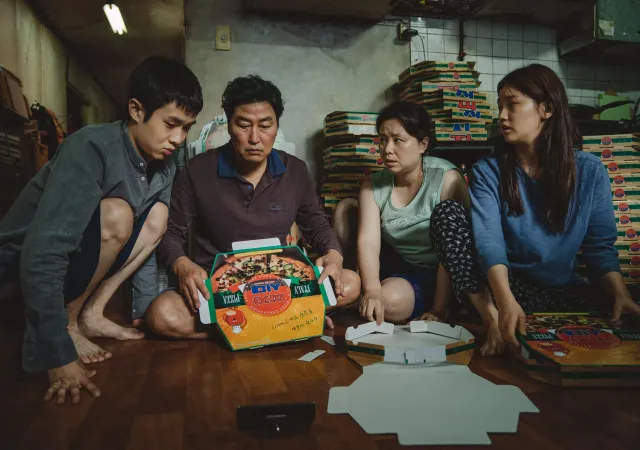 Four Korean family members sit on the floor in a cramped room surrounded by stacks of pizza boxes. The father holds a pizza box while the others look at it with serious, concerned expressions. An unfolded pizza box lies on the floor in front of them.