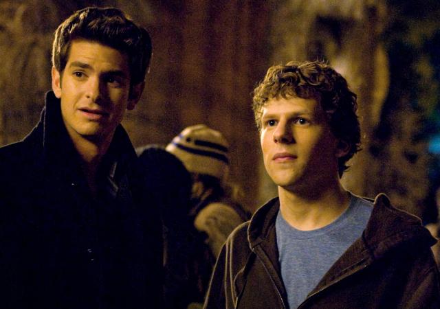 Two young men stand side by side in a dimly lit outdoor setting at night, surrounded by a crowd. One wears a dark coat, while the other wears a blue T-shirt under a brown hoodie. Both look off-camera with serious, attentive expressions, illuminated by warm golden lighting.