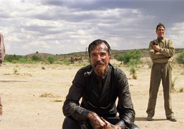 A mustached man in a dark suit crouches in the foreground of a dry, open landscape with a pipe in his mouth. Two men stand behind him on either side, one wearing a cowboy hat and the other with his arms crossed. Sparse vegetation, small buildings, and a cloudy sky stretch across the background.