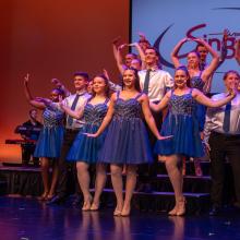 Wisconsin Singers pose on the stairs wearing blue sparkly dresses and silver shirts with an orange backdrop behind them.
