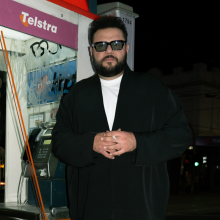 Photo of Mo Amer dressed in black, standing against a phone booth.