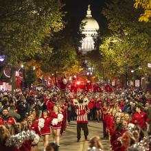 Bucky Badger struts down State Street with cheerleaders and marching band members as audience members cheer them on from the parade sidelines.