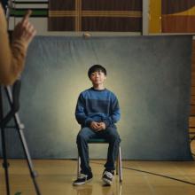 Still from film Didi, kid sits in front of grey background for school picture day.