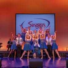 A group of performers in blue shimmery dresses and collared shirts and ties pose in front of a banner that reads "Wisconsin Singers" while band members play various instruments around them.