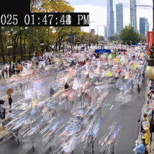 An image from a camera looking at a busy street in a non-descript city. There is a timestamp in white text over a black box in the upper left-hand corner. People are moving quickly and are blurred due to the motion. The image is slightly grainy as it is taken from a surveillance camera. There are trees on the opposite side of the street, and buildings and lampposts on the side of the street the camera is on.a 