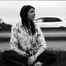 A black-and-white image of artist Anne E. Stoner. She is sitting down near the side of a road, with cars in motion behind the guardrail behind her. She is holding a microphone to record the sounds of traffic. She is looking to the right side of the image. She has long dark hair and is wearing light-colored shirt with a light pattern.
