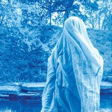 A photograph that is entirely Prussian blue in color, of a figure draped in a cloth or bedsheet to appear like a ghost, as the sheet seems to be blowing in the wind. The image is taken outside, with a natural background featuring trees and a hillside.