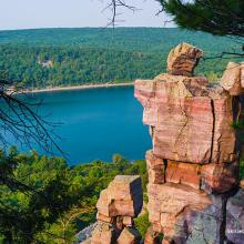Photo of Devil's Lake landscape showing big boulders and rock stacked on edge of cliff with view of lake and greenery.