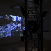 A dark image from inside Anne Stoner's Infrastructure Bodies/Injury Systems. In the foreground there is a metal structure with various speaker and wires strewn about. Behind the structure on the wall is a projected image of a streetscape with people and cars, the image is "datamoshed" meaning that pixels are merging into one another. The windows of Main Gallery are slightly visible in the background.