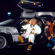 two individuals, one seated in a DeLorean vehicle and one crouching next to the vehicle