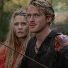 woman in red standing to the left of man in black holding a thin sword
