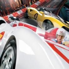 White race car with red trim and a helmeted driver, followed by a yellow race car.