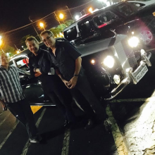 Three men stand in front of an old car, smiling for the camera.