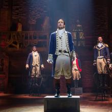 Image showing several people on stage.  Front and center is actor Lin-Manuel Miranda as Alexander Hamilton.