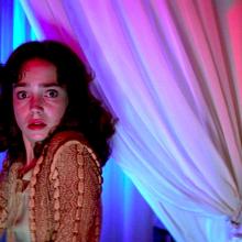 woman stands in a curtain-covered room holding a knife looking shocked and scared